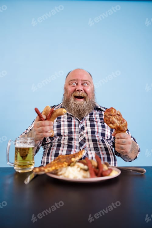 Preview: Excited obese man holds chicken leg and bunch of sausages at table in studio