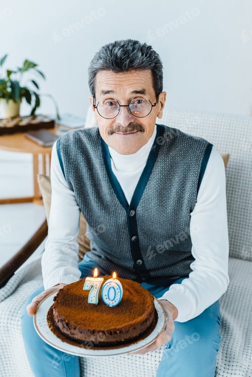 Preview: happy senior man holding birthday cake while sitting on sofa a in living room