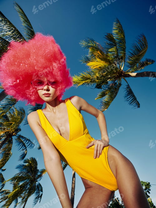 Preview: Bright yellow swimsuit, pink wig, woman posing near tropical palm trees, vibrant setting, sunny day