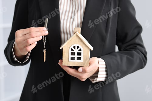 Preview: Real estate agent holding key with keychain and house model indoors, closeup