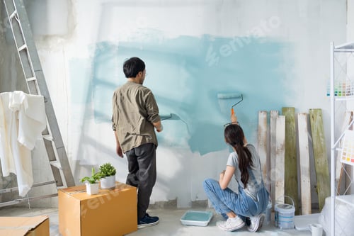 Preview: Asian couple repairing and painting the wall with blue paint using a roller during renovation