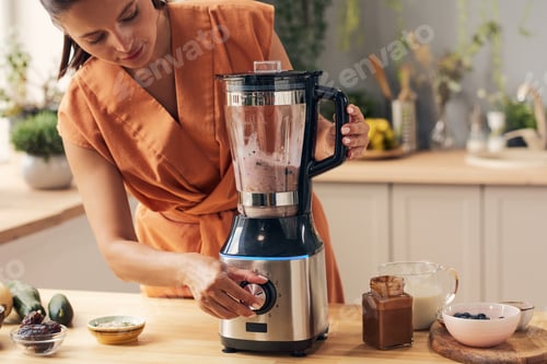 Preview: Young female mixing ingredients in electric blender while making smoothie