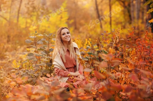 Preview: Beautiful young blonde woman in dress sitting with pensive look in tall grass in autumn forest