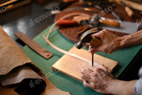 Preview: Male artisan holding a tool and a hammer to puncture or shape the leather on a workbench