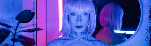 Preview: Portrait of drag queen in wig looking at camera in neon lighting at home, banner