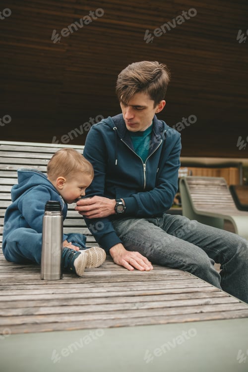 Preview: child and dad are sitting on a deck chair on the river bank in spring and drinking tea from thermos