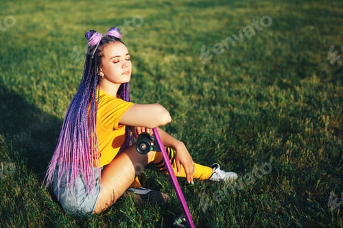 Preview: Young beautiful girl sitting on a lawn holding a skateboard in her hand in a yellow T-shirt and