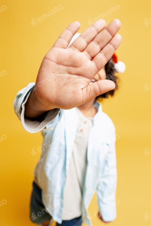 Preview: Shy child hiding face with hand, wearing Santa hat on yellow background