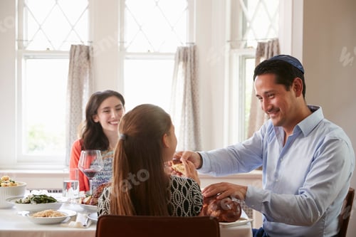 Preview: Jewish man sharing challah bread with family at Shabbat meal