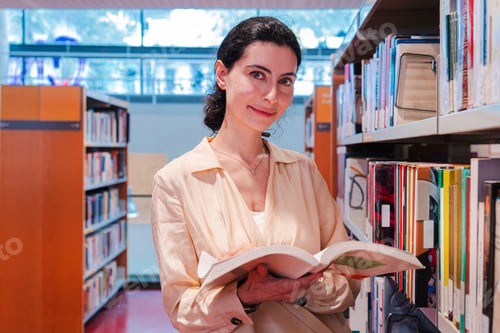 Preview: Charming Woman Smiling While Holding an Open Book in a University Campus Library Filled with Diverse
