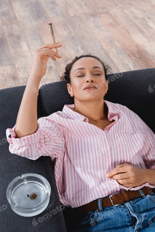 Preview: high angle view of african american woman lying on sofa and smoking rolled joint with medical