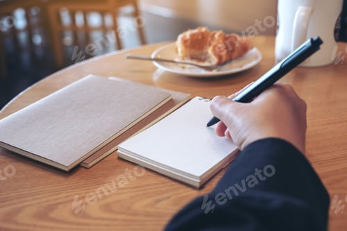 Preview: Closeup image of a hand writing on blank notebook with coffee cup and croissant on table in cafe