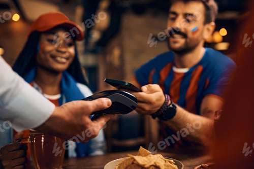 Preview: Close up of couple of sports fans using smart phone and paying contactless in bar.
