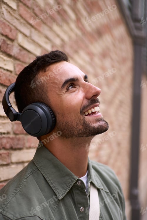 Preview: Close up handsome young man looking up smiling with wireless headphones leaning against brick wall.