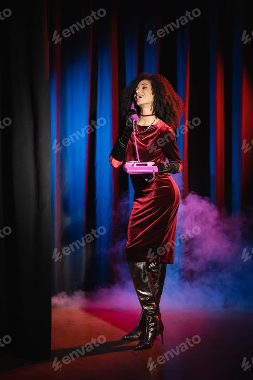 Preview: Cheerful african american woman in velvet dress talking on telephone on blue background with light