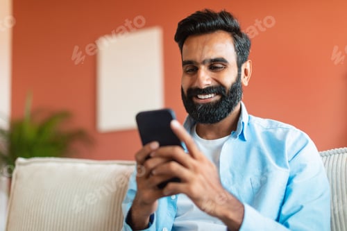 Preview: Cheerful Indian Man Using Mobile Phone Surfing Internet Sitting Indoor
