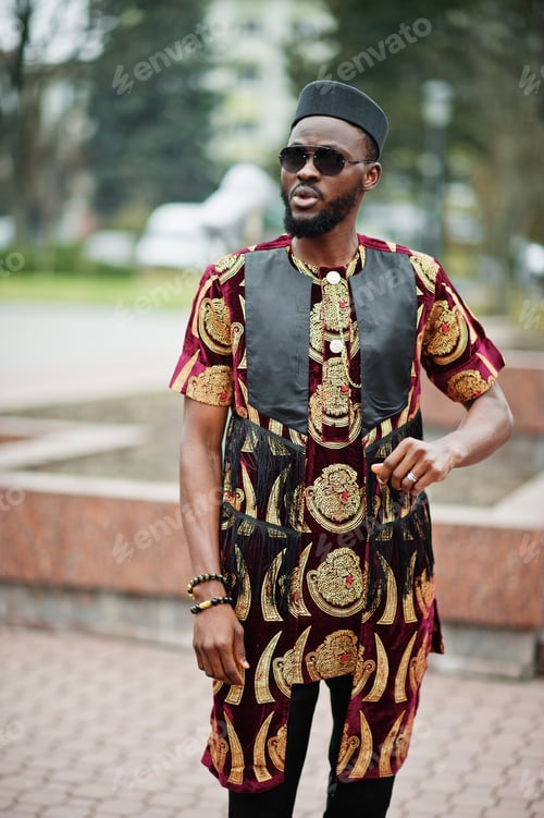 Preview: African stylish and handsome man in traditional outfit and black cap standing outdoor.