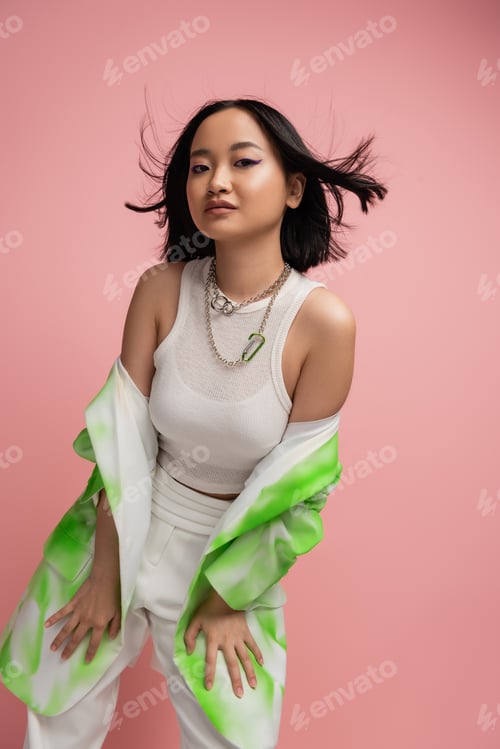 Preview: trendy asian woman in tank top and green and white blazer posing on wind isolated on pink