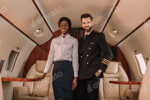 Preview: handsome, positive pilot standing with hand in pocket near smiling african american stewardess in