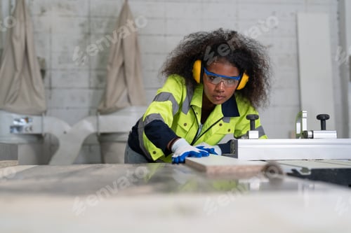 Preview: Black female carpenter wear ear protection glasses safety use circular saw cutting wood at factory