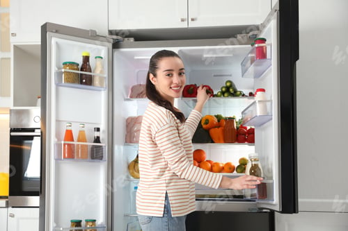 Preview: Young woman taking bell pepper out of refrigerator in kitchen