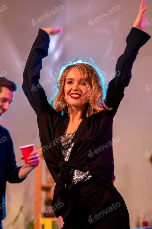 Preview: Young cheerful woman with toothy smile and raised arms dancing at home party