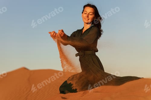 Preview: woman in green long dress stands in desert sand dunes background yellow orange sunset