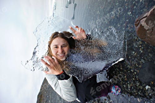 Preview: Cheerful pretty young woman looking through a large piece of ice on a rocky shore