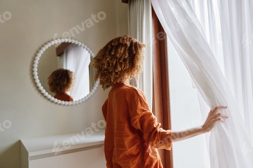 Preview: A young woman with curly hair stands by a window, gently pulling back a sheer curtain in a cozy