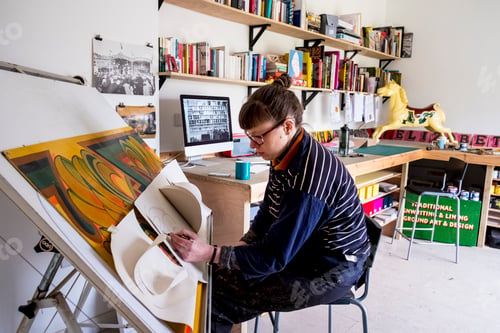 Preview: Woman artist sitting at a tilted drawing table in her studio, drawing, using several sheets of stiff