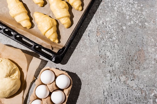Preview: Top View of Uncooked Croissants on Baking Tray Near Eggs And Dough on Concrete Grey Surface