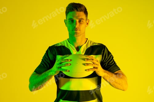 Preview: Portrait of caucasian male rugby player with rugby ball over yellow lighting