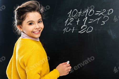 Preview: cheerful schoolgirl in yellow holding chalk and looking at camera in front of blackboard