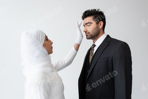 Preview: young bride in hijab touching forehead of groom during wedding ceremony isolated on grey