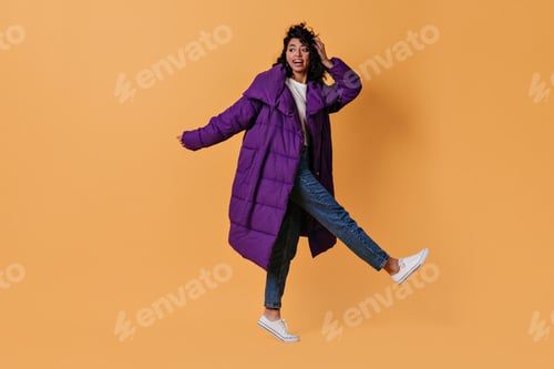 Preview: Amazed mixed race girl in down jacket standing on one leg. Full length view of dancing curly woman