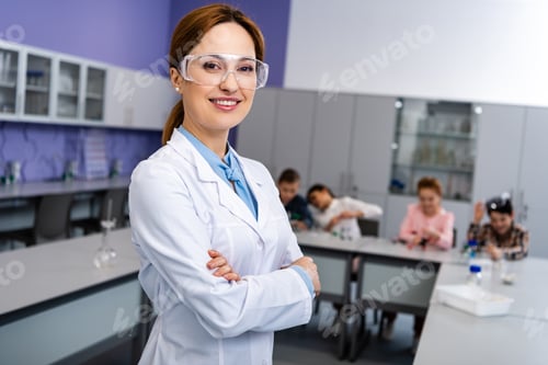 Preview: Smiling teacher in protective goggles standing with crossed arms in front of pupils during chemistry