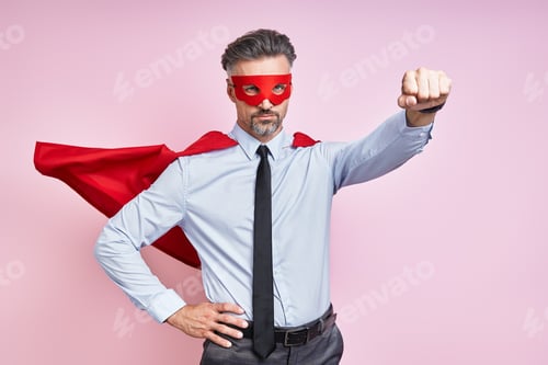 Preview: Confident mature man wearing superhero cape and keeping arm outstretched against pink background