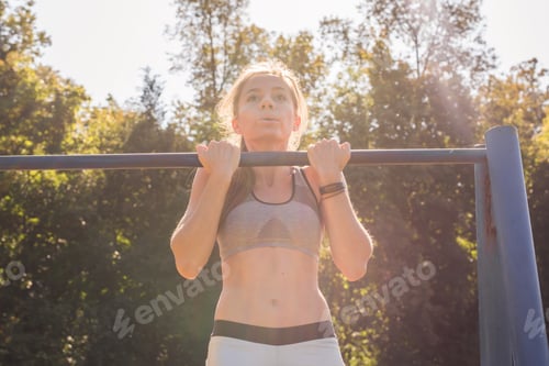 Preview: young athletic fitness woman working out at outdoor gym doing pull ups at sunrise