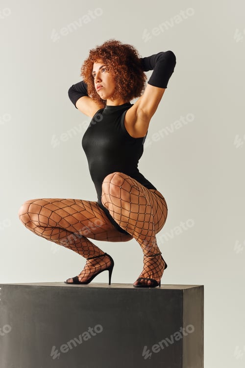 Preview: appealing young african american woman in sexy black attire squatting on black cube, fashion concept