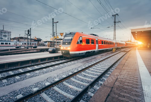 Preview: Railway station with beautiful modern red commuter train at suns