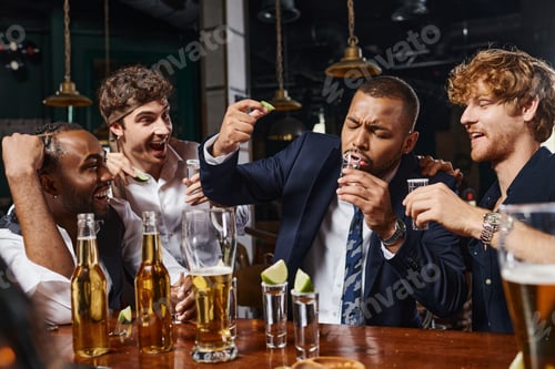 Preview: happy interracial men looking at african american friend drinking tequila shot and holding lime