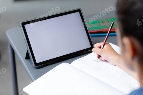 Preview: Biracial schoolgirl sitting at desk writing in class and using tablet, with copy space on screen