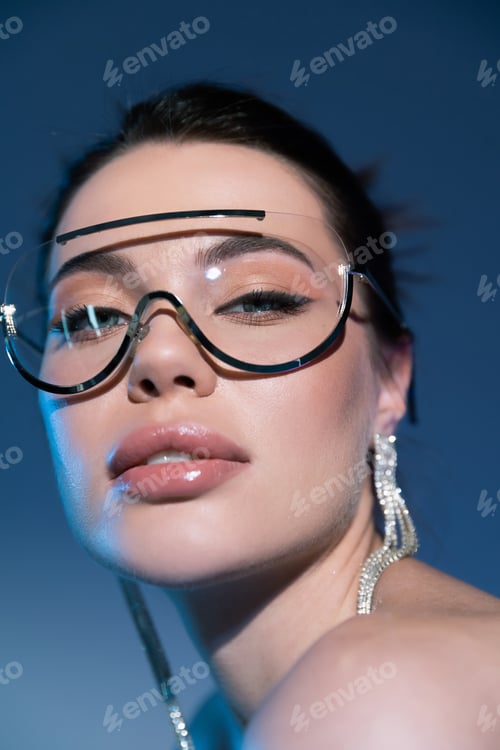 Preview: close up portrait of woman in makeup and stylish sunglasses looking at camera on blue background
