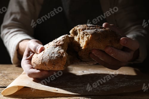 Preview: Man breaking loaf of fresh bread at wooden table, closeup