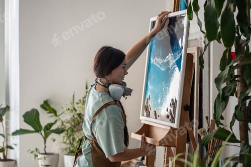 Preview: Focused artistic woman putting canvas with epoxy resin painting in liquid art technique on easel.