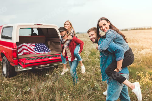 Vista previa: young happy women piggybacking on boyfriends in flower field during car trip