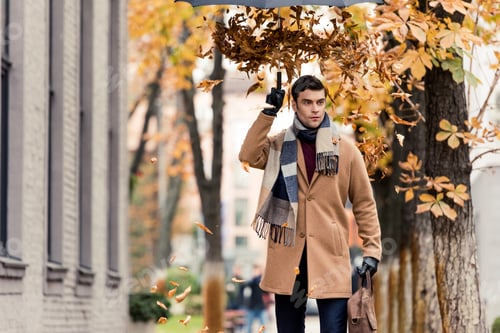 Preview: stylish man in coat with golden leaves falling from umbrella standing on autumnal street