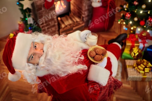 Preview: Top view. Santa Claus with milk and cookies in hands looks upward while sitting in a chair