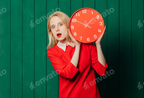 Preview: Surprised blonde hair woman in red dress with clock on green wooden background