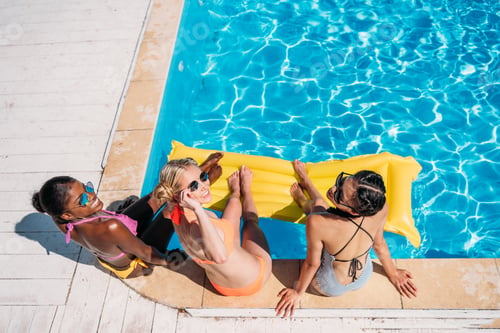 Preview: Young beautiful multiethnic women sitting near swimming pool at resort